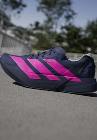 Chaussure de course bleu foncé avec des rayures rose vif et une semelle "Lightstrike Pro" placée sur une surface pavée en extérieur.