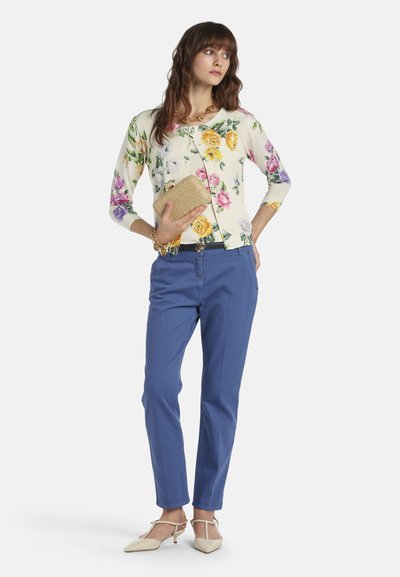 Woman wearing a floral cardigan, blue trousers, beige heels, gold necklace, and bracelet, holding a woven clutch purse.