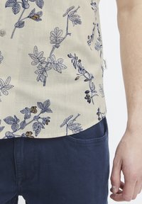 Camisa de manga corta beige con estampado floral en navy, que presenta hojas y flores detalladas. Combinada con pantalones azul marino.