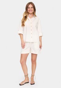 Light beige button-up shirt with rolled sleeves and matching shorts, featuring an elastic waistband and cuffed hems. Flat sandals complete the look.