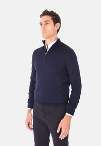 Maglione zip-up blu navy con colletto alto, realizzato in un materiale a maglia liscia, abbinato a pantaloni neri. Presenta un'accortezza bianca sul colletto interno.