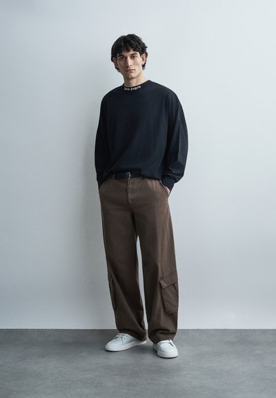 Giovane uomo che indossa una maglietta a maniche lunghe nera, pantaloni cargo marroni, sneakers bianche, in piedi contro un muro grigio chiaro uniforme.