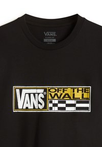 Svart bomull t-shirt med en framträdande vit och gul Vans-logotyp med texten "OFF THE WALL," accentuerad av ett rutmönster till höger.