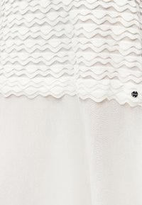 Άσπρο ύφασμα με οριζόντιες φουσκωτές κυματιστές πτυχώσεις στο άνω τμήμα και λείο ημιδιάφανο υλικό από κάτω, με μικρό στρογγυλό μεταλλικό ταμπελάκι.