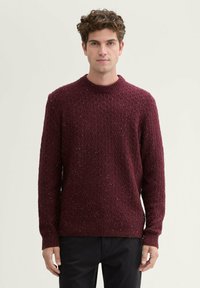 Burgunderfarbener Strickpullover mit einem strukturierten Muster, geripptem Rundhalsausschnitt und Bündchen, der dezente funkelnde Akzente aufweist. Schwarze Hose.