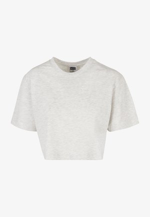 Frau trägt ein lockeres, kurzärmeliges, hellbeiges Cropped-T-Shirt und eng anliegende schwarze Hosen vor einem schlichten grauen Hintergrund.