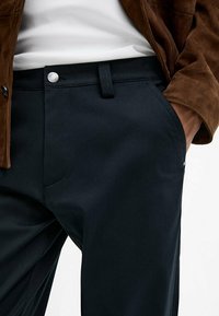 Navy trousers made from smooth material, featuring a flat front, belt loops, and a silver button accent. Considered versatile and modern design.