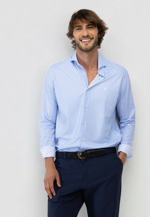 Hombre sonriendo, vestido con una camisa de cuadros azul claro con puños remangados y pantalones azul oscuro, de pie frente a un fondo claro y sencillo.