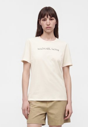 Žena s rovnými hnedými vlasmi, oblečená v krémovom tričku Michael Kors a béžových šortkách, stojaca pred jednoduchým pozadím.