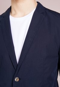 Granatowa, lekka marynarka zapinana na jeden guzik, z klapami o kroju notch oraz gładką fakturą, noszona na białej koszulce.