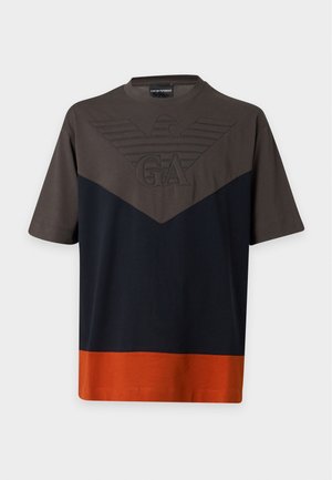 Νέος άνδρας που φοράει ένα t-shirt με χρωματική μπλοκ σε καφέ, μαύρο και πορτοκαλί, ανοιχτό μπλε φαρδιά τζιν και μαύρα παπούτσια, στέκεται με το ένα χέρι στην τσέπη.