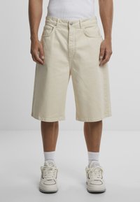 Beige denim shorts with a loose fit, featuring a button fly, five pockets, and a straight hem. Paired with white sneakers and socks.