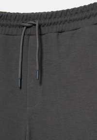 Gros plan sur la ceinture d’un pantalon de survêtement gris foncé avec bande élastique et cordon noué avec embouts en plastique.