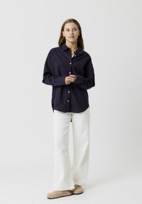 Camicia blu navy a maniche lunghe con righe rosse, abbinata a pantaloni bianchi a gamba larga e scarpe beige slip-on. Collo semplice e vestibilità rilassata.