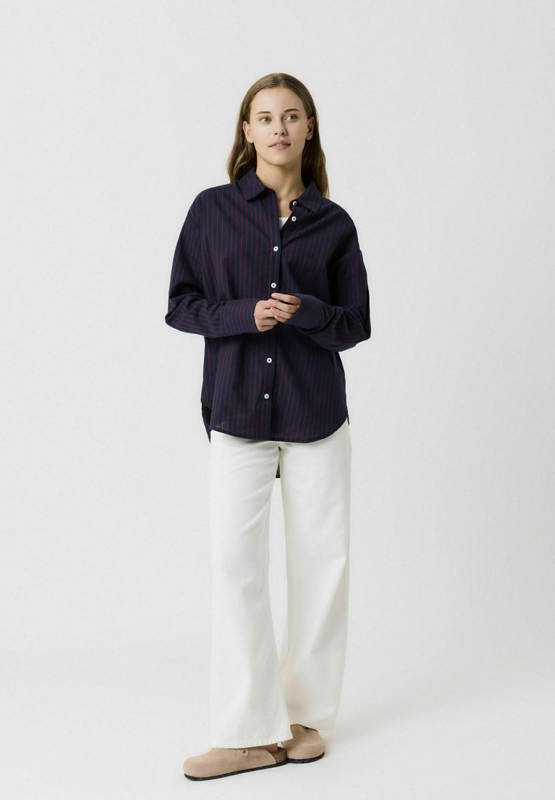Camicia blu navy a maniche lunghe con righe rosse, abbinata a pantaloni bianchi a gamba larga e scarpe beige slip-on. Collo semplice e vestibilità rilassata.