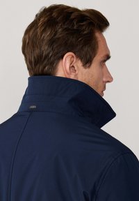 Man met bruin haar die naar rechts kijkt, gekleed in een marineblauwe jas met een hoge kraag en een klein Hackett London-logo op de rug.