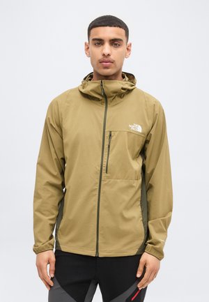 Jeune homme portant une veste à capuche couleur beige de The North Face avec une poche zippée sur la poitrine, debout devant un fond clair uni.
