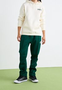 Sweat à capuche crème avec poche avant et logo, associé à un jogging vert foncé avec un patch logo. Baskets montantes noires avec semelles blanches.