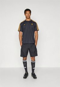 Μαύρη ριγέ κοντομάνικη φανέλα με κίτρινες λεπτομέρειες, συνδυασμένη με μαύρο σορτς και μαύρες κάλτσες μέχρι το γόνατο με λευκές ρίγες.