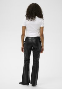 Juoda odinė išplatėjusi kelnės su lygiu paviršiumi, aukštu juosmeniu ir užpakalinėmis kišenėmis, vilkima su prigludusia balta marškinėliais. Matoma iš nugaros.