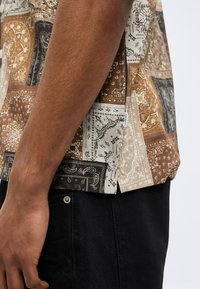 Χέρι και πλευρό άνδρα που φοράει καφέ, μπεζ και μαύρο πουκάμισο με κοντά μανίκια και μαύρο παντελόνι, μπροστά σε απλό φόντο.