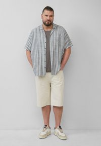 Kortebloemige gestreepte shirt in blauw en wit, grijze T-shirt, lichtbeige shorts en sneakers met tan accenten. Staand tegen een neutrale muur.