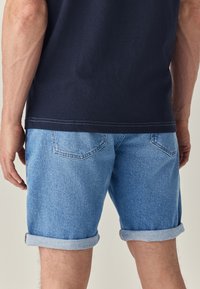 Shorts in denim blu con risvolti, dotati di tasche posteriori e finitura testurizzata. Indossati con una camicia di colore scuro, che mostra uno stile casual.