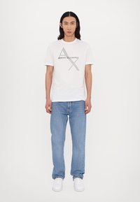 Fehér pamut póló az "ARMANI EXCHANGE" logóval és geometrikus mintával. Világoskék farmernadrággal és fehér sportcipővel párosítva.