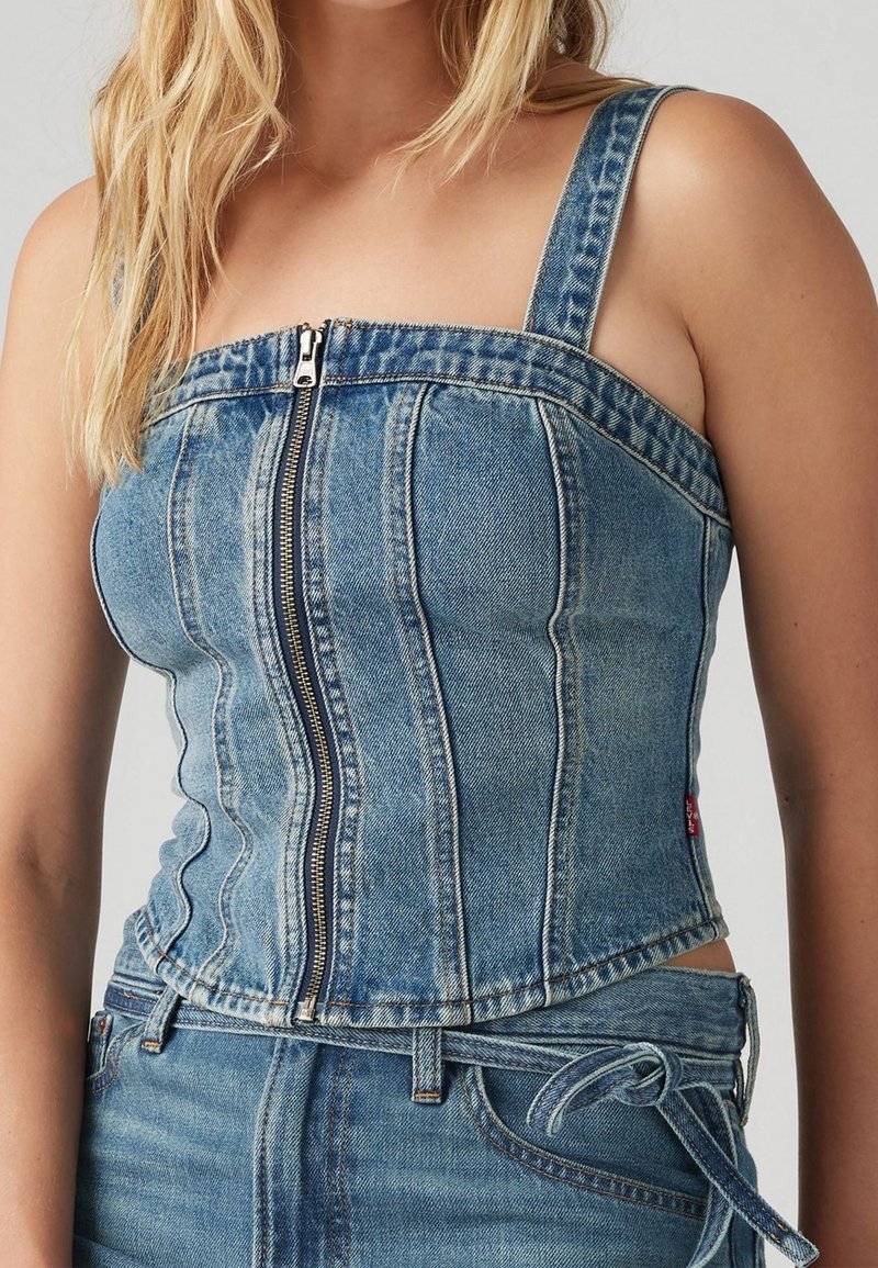 Kvinna i blå denim korsettdesign topp med framzipper och matchande denimjeans med knytbälte, visad från halsen till höfterna.