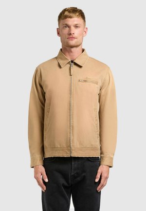 Mann trägt eine hellbraune Reißverschlussjacke mit Kragen und Brusttasche, kombiniert mit schwarzen Hosen, steht vor einem schlichten hellen Hintergrund.