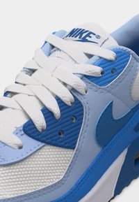 Κοντινό πλάνο ενός μπλε και άσπρου αθλητικού παπουτσιού Nike με λευκά κορδόνια, υφασμάτινο πλέγμα και το λογότυπο Nike swoosh στην πλευρά.