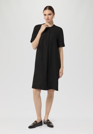 Schwarzes, kurzärmliges Kleid aus strukturiertem Stoff, mit rundem Halsausschnitt, Knopfverschluss und lockerer Passform. Kombiniert mit schwarzen Mokassins.