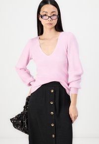 Suéter cor-de-rosa canelado com decote em V e mangas longas, combinado com uma saia preta com detalhes em botão e uma bolsa floral.