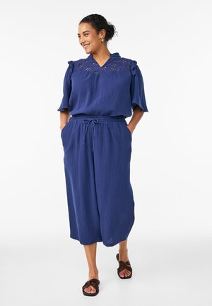 Smilende kvinde med mørkt hår i en knold, iført en blå tekstureret bluse med blondeindlæg og brede, korte bukser, brune sandaler, hænderne i lommerne.