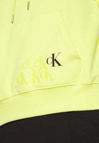 Jasnożółta kieszeń z kapturem z powtarzającym się logo "cK" w różnych odcieniach, połączona z czarnymi spodniami.