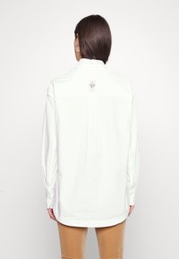 Kvinna med rakt brunt hår som bär en vit långärmad skjorta med texten "50 år" på övre ryggen och kamelfärgade byxor.