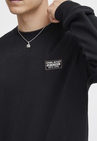 Svart sweatshirt med rund halsringning, ribbade muddar och en liten märkespatch på bröstet. Utrustad med ett silverkedjehalsband med ett hänge.