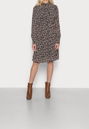 Schwarzes Blumenkleid mit langen Ärmeln und hohem Ausschnitt, verziert mit kleinen weißen und orangefarbenen Blumen. Kombiniert mit braunen Knöchelstiefeln.