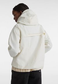 Let lightvægter hvid jakke med hætte, tekstureret stof og en baglomme med gyldne detaljer. Båret over en mønstret beige skjorte og sorte bukser.