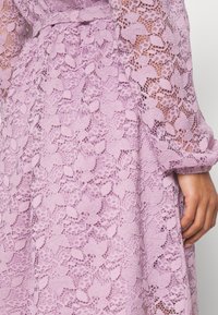 Gros plan d'une main reposant sur une robe en dentelle florale lavande avec des manches longues transparentes et des motifs complexes de feuilles.