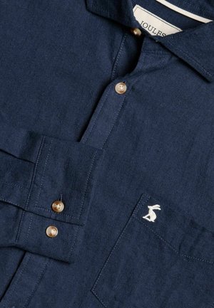 Laivansinisestä pellavapaita, jossa on napitettava kaulus. Paidassa on kultaisia nappeja ja valkoinen brodeerattu pupulogo rintataskussa.