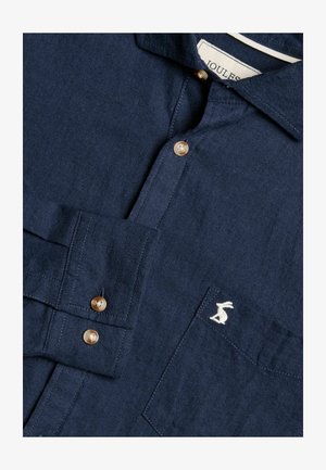 Laivansinisestä pellavapaita, jossa on napitettava kaulus. Paidassa on kultaisia nappeja ja valkoinen brodeerattu pupulogo rintataskussa.