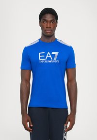 Blue short-sleeve t-shirt featuring the logo "EA7 EMPORIO ARMANI" in white. Striped accents on shoulders, smooth fabric, fitted design.
