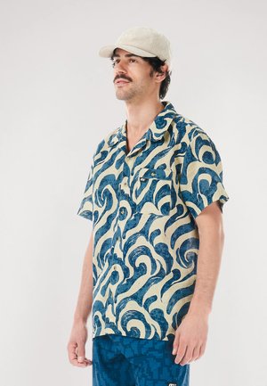 Kurzärmeliges Hemd in Creme und Blau mit Wellendesign; verfügt über eine aufgesetzte Tasche und einen Kragen; kombiniert mit gemusterten blauen Shorts.