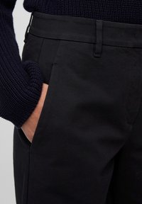 Hand in de zak van zwarte op maat gemaakte broek, gedragen met een donkerblauwe grofgebreide trui.