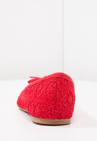 Ballerine rosse in pizzo con motivi floreali, punta arrotondata e suola beige, viste da dietro su una superficie bianca.