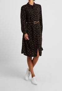 Czarna sukienka z długimi rękawami i pomarańczowym wzorem w klucze, z rozcięciem z przodu i błyszczącym paskiem, w połączeniu z białymi trampkami.