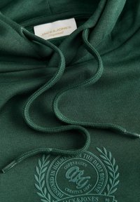 Sweat à capuche vert foncé avec cordons et motif circulaire embossé avec texte et branches de laurier, étiquette de la marque visible à l'intérieur du col.