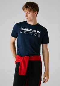 Navyblaues Baumwoll-T-Shirt mit einem weißen "Red Bull Racing"-Logo und zwei roten Stieren. Kombiniert mit einem roten Sweatshirt, das um die Taille gebunden ist.