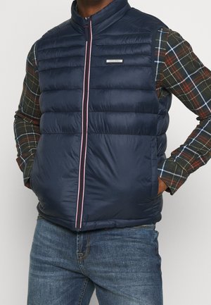 Homme portant une veste matelassée bleu marine sur une chemise à carreaux verts et rouges, les mains dans les poches de la veste, associée à un jean bleu.